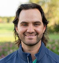 Head and shoulders photograph of a smiling Noah Cronin, Program Coordinator of Manatee County, Level 2 Coach and TPI Certified Level 1 of First Tee – Sarasota/Manatee standing proudly in front of a golf course.